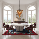 A luxury dining room featuring a round white marble dining table surrounded by six dark wood chairs with burgundy upholstery. The room is decorated with a large gold-framed octagonal mirror, a multi-tiered crystal chandelier, and a red and blue patterned rug on a light stone floor.