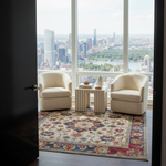 A styled interior view from a dark doorway into a modern living room with two cream boucle armchairs, a striped side table, and a multicolor patterned area rug, all overlooking a city skyline and river through large floor-to-ceiling windows.