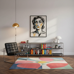 A modern living room scene featuring a colorful abstract area rug on a herringbone wood floor, a black leather armchair, a metal shelving unit with books and decor, and a framed portrait print on the wall.