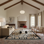 A styled living room interior with a beige sofa, two wooden armchairs, a white round coffee table, a large floral area rug, a brick fireplace, and a white pendant light in a bright, high-ceilinged room.