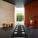 A modern interior scene with a long black runner rug featuring a geometric pattern of white and light beige rectangles, placed on a dark floor between two curved wooden walls.