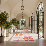 A long arched corridor with cream walls, hanging lantern lights, large black-framed glass doors, potted orange trees, and several rectangular area rugs featuring large pink and orange floral motifs on a light background.