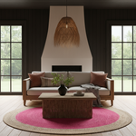 A styled living room interior featuring a light wood sofa with neutral and rust-colored cushions, a woven rattan coffee table, a round pink and beige rug, dark wood paneled walls, and a large woven pendant light hanging above a fireplace.