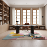 A large, colorful abstract art rug with a stylized face design is displayed in a modern, sunlit home office. The rug features a vibrant mix of pastel blue, pink, yellow, and cream, with a bold graphic face motif. The room is styled with light wood flooring, a matching wood desk, a brown leather chair, and built-in wooden bookshelves in the background.