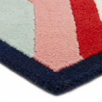 Close-up of a textured rug corner showing bold color-blocked stripes in pale mint, pink, red, and dark navy on a white background.