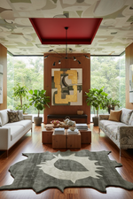 A modern living room with a large abstract art print hanging on a wood-paneled wall, flanked by potted plants and sofas on both sides. The room features a patterned ceiling, a red recessed light, hardwood floors, and a large organic-shaped rug in the foreground.