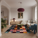 A styled living room interior with a colorful patchwork area rug on a light wood herringbone floor, a cream sectional sofa, a green coffee table, a blue accent chair, and a burgundy pendant light.