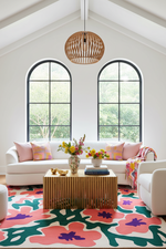 A bright, modern living room with a white sectional sofa, pink accent pillows, a slatted wood coffee table, a colorful floral area rug, and a woven pendant light hanging from a high vaulted ceiling.