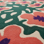 A close-up of a textured area rug with a bold, organic floral pattern in coral pink, dark green, cream, and purple.