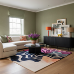 A styled living room interior with sage green walls, a cream sectional sofa, a black sideboard with framed art and lamps, a colorful abstract area rug, and a round black coffee table with a vase of purple flowers.