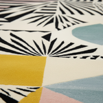 Close-up of a modern geometric area rug with a cream base, bold black fan-like stripes, a large mustard yellow block, a pale blue shape, and a soft pink section.
