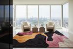 A modern living room with two cream armchairs, two small side tables, and a large abstract area rug in black, pink, mustard yellow, and beige. The room is positioned in front of floor-to-ceiling windows overlooking a city skyline and Central Park.