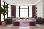 A modern living room featuring two gray fabric sofas facing a low glass coffee table, with two round mauve ottomans on a pink and lavender scalloped area rug. The room has a central floor-to-ceiling window with dark brown drapes, a built-in fireplace, and a large potted plant in a gold planter.