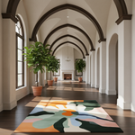 A large abstract area rug with a multicolor floral pattern in orange, cream, sage green, pink, and dark green, displayed on a dark wood floor in a bright, classic hallway with arched windows and potted plants.