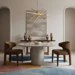 A modern dining room scene featuring a round marble-top table with a textured stone pedestal base, four upholstered dining chairs in brown, a gold starburst chandelier, and a colorful abstract area rug.