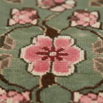 Close-up of a woven floral rug with a large pink flower and smaller blossoms on a muted sage green background.