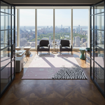 A modern living room interior with a large pink and white patterned area rug on a wood floor, two black armchairs, and expansive floor-to-ceiling windows overlooking a city skyline.