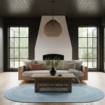 A styled living room interior featuring a light wood sofa with neutral and rust accent pillows, a round light blue rug, a wooden coffee table, and a large woven pendant light hanging in front of a white fireplace with dark wood trim.