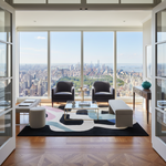 A luxury modern living room with a panoramic view of the New York City skyline from floor-to-ceiling windows. The room features two dark gray armchairs, a glass-top coffee table, a black and multicolor abstract area rug, a long beige bench, and a wooden herringbone floor.