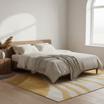 A minimalist bedroom scene featuring a low wooden platform bed dressed in cream bedding and a textured gray throw blanket, set on a modern abstract yellow, cream, and gray patterned area rug. The room has light wood floors, a matching wooden headboard, a small side table with a vase of dried grass, and a bright window on the right.