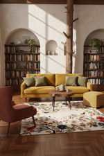 A styled living room scene featuring a mustard yellow sofa with olive green pillows, a rust-colored armchair, a patterned floral area rug, and a round wooden coffee table. The room has a textured off-white wall with arched alcoves filled with bookshelves, a central tree trunk, and warm sunlight streaming in.