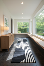 A modern hallway or entryway with a long black and white striped runner rug on dark floor tiles. The space features a light wood credenza, a table lamp, built-in window benches, and large windows looking out to green foliage.