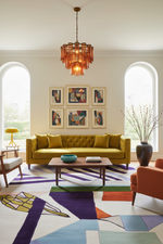 A modern living room featuring a mustard yellow tufted sofa, a geometric multicolor area rug, a wooden coffee table, and a copper-toned chandelier hanging from the ceiling.