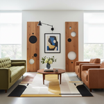 A modern living room interior with a green sofa on the left, two tan leather armchairs on the right, a round wooden coffee table, a patterned area rug, and a framed abstract artwork centered on a white wall between two tall wooden panels with circular cutouts.