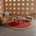 A styled living room scene featuring a round woven area rug with a red center and natural jute border, surrounding a wooden coffee table. The room has a terracotta-colored wall with circular cutouts, a curved tan sofa, and a rattan armchair.