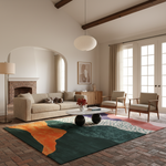 A modern living room with a large abstract area rug in green, orange, and teal tones, styled with a beige sofa, two wooden armchairs, a round coffee table, and a brick fireplace in the background.