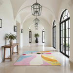 A colorful abstract area rug with pastel pink, blue, yellow, and cream sections is displayed on a light stone floor in a bright, elegant hallway with arched ceilings, hanging lanterns, and large black-framed windows.