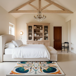 A styled bedroom interior with a cream upholstered bed, white bedding, a tall light wood bookcase, a dark wood door, a hanging chandelier, and a colorful abstract rug on the floor.