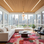 A modern living room with a white sectional sofa, a dark wood coffee table, black armchairs, and a bold floral area rug in pink, red, and beige tones. The room is set in a sunlit balcony with a wood-paneled ceiling, linear LED lights, and a large window offering a city skyline view.