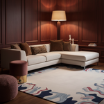 A modern L-shaped sectional sofa in off-white fabric with brown and leopard-print throw pillows, styled in a luxurious room with dark wood paneling, a floor lamp, and a large abstract-patterned rug.
