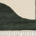 Close-up of a textured woven rug corner showing a dark forest green section, a warm cream section, and cream fringe along the bottom edge.
