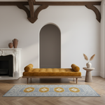 A styled interior scene showing a mustard yellow tufted daybed centered in a room with a white fireplace, arched wall opening, wooden ceiling beams, a small side table with a vase of dried grass, and a blue patterned area rug on a wood floor.