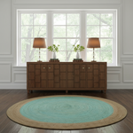 A round area rug with a turquoise center and natural jute border, styled on a dark wood floor in front of a mid-century modern wooden credenza with lamps and plants.
