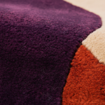 Close-up of a textured area rug with a color-blocked design, featuring a large deep purple section, a warm rust-orange section, and a cream section.