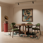 A styled dining room scene featuring a round wooden table and four green upholstered chairs on a floral rug, with a framed abstract painting on the wall and a vintage gramophone on a red pedestal.