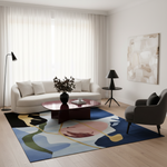 A modern living room featuring a large abstract area rug with blue, beige, green, and pink shapes. The room includes a cream-colored sofa, a dark wood coffee table, a gray armchair, black floor lamps, and a large abstract canvas painting on the wall.