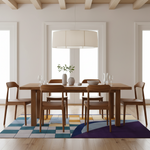 A modern dining room scene with a rectangular wooden dining table and four matching wooden chairs arranged on a colorful geometric area rug. The room has light wood floors, white walls, large windows, and a large drum pendant light overhead.