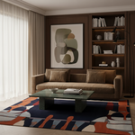 A styled living room interior featuring a brown fabric sofa, a green glass coffee table, a large abstract wall art print, and a bold geometric area rug in navy, orange, beige, and gray tones.