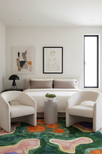 A modern bedroom interior with a bed, two cream armchairs, framed wall art, a small side table, and a bold abstract patterned rug in green, pink, orange, and purple tones.
