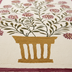 Close-up of a woven textile or rug featuring a gold pot with olive green leafy stems and burgundy flowers on an off-white background.