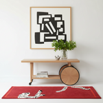 A styled interior scene with a red area rug featuring black-and-white dalmatian spots, placed beneath a light wood console table and a framed abstract black-and-white artwork on a white wall.