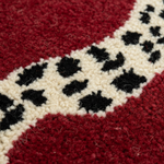 Close-up of a textured rug with a deep red field, a cream border, and black abstract spots.