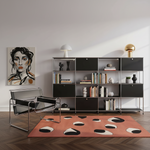 A modern living room scene with a large black and white geometric patterned area rug on a dark wood floor, a black metal lounge chair, a black shelving unit with books and decor, and a framed portrait painting on the wall.