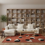 A modern living room setup featuring a large white sectional sofa, a light wood coffee table, a rust-orange area rug with black and cream abstract spots, and a massive light wood bookshelf filled with books and decorative objects in the background.