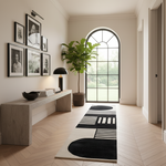 A modern interior hallway with a long rectangular area rug featuring bold black and white geometric shapes, a light wood console table, framed black-and-white portraits on the wall, and a large potted plant near an arched window.