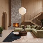 A modern living room with a large olive green sectional sofa, a round wooden coffee table, a black and beige area rug, a stone fireplace with a glowing fire, and a wooden staircase in the background.